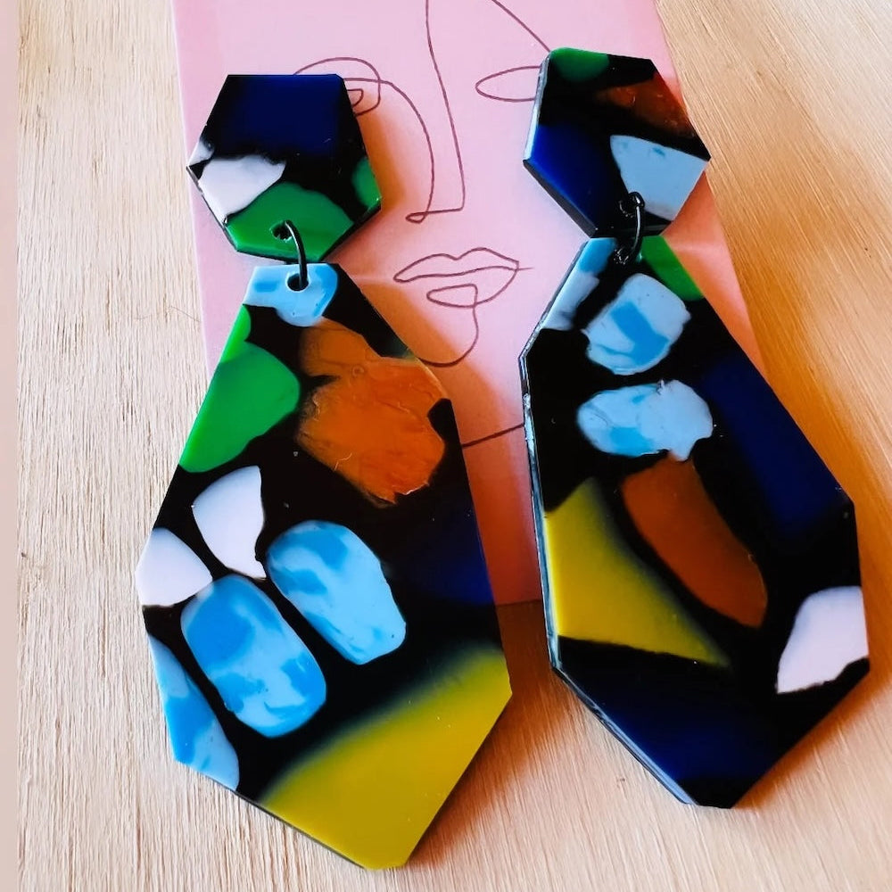 Colourful abstract earrings made from recycled plastic on a cardboard backing sitting on a wooden table.