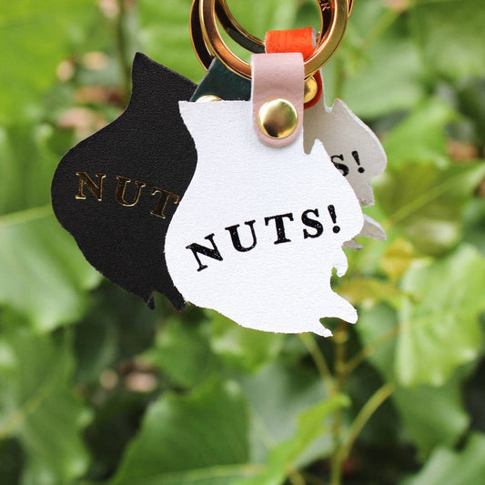 Keychain with black and white squirrel-shaped tags reading 'NUTS!' against a green leafy background