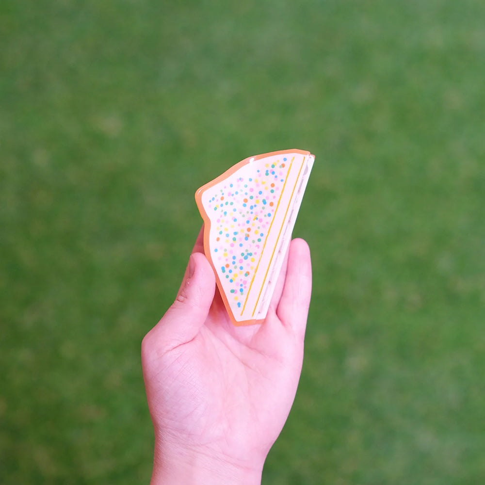 Hand holding a hair claw in the shape of fairy bread against a blurred green background.