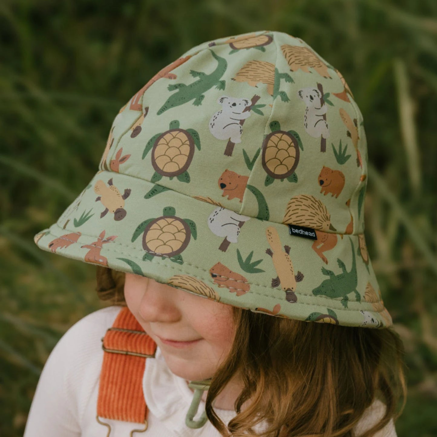 Side view of toddler wearing an Australian animal patterned bucket hat against a blurred green background