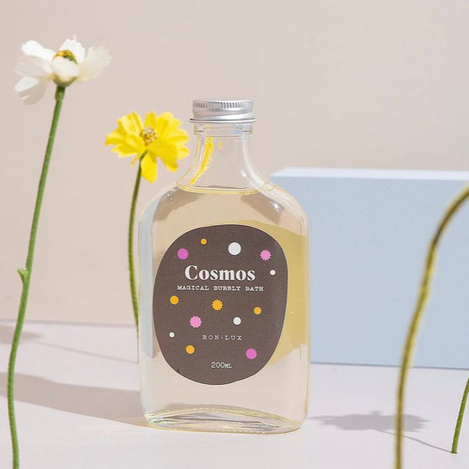 Bon Lux Cosmos Magical Bubbly Bath in a glass bottle, styled with yellow and white flowers on a white background.