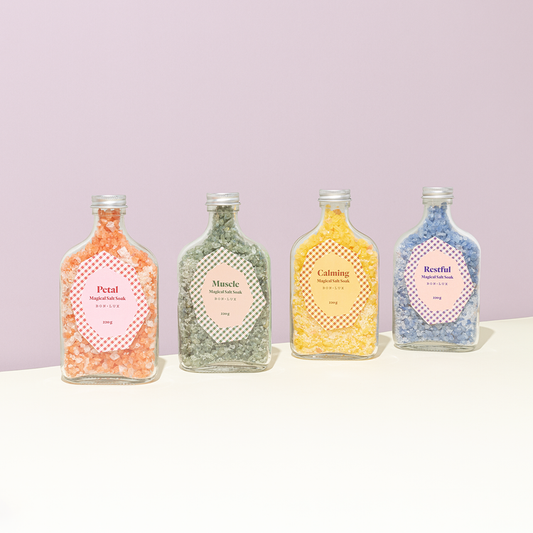 Four Bon Lux Magical Salt Soak bottles with colourful contents on a white surface against a lilac background.