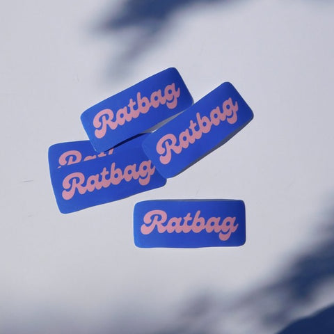 Flat lay of some Carla Adams Ratbag Vinyl Sticker. Sticker text is in a bubbly italic font in light pink and the background is in bright blue.  