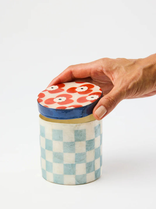 Hand holding a ceramic jar with a checkered pattern and floral lid on a white background