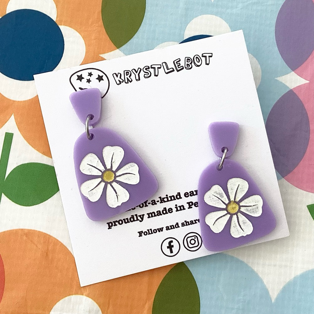 Purple earrings with white flowers on a Krystlebot card against a colourful background.