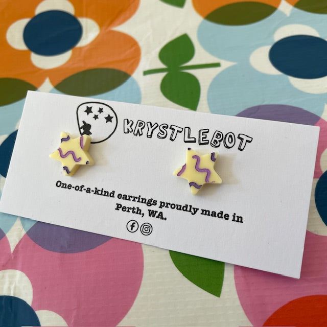Krystlebot Squiggle Star Studs - Cream/Lilac/Purple. Star-shaped earrings with a cream coloured base and lilac and purple squiggles on a 'KRYSTLEBOT' branded card with colorful background