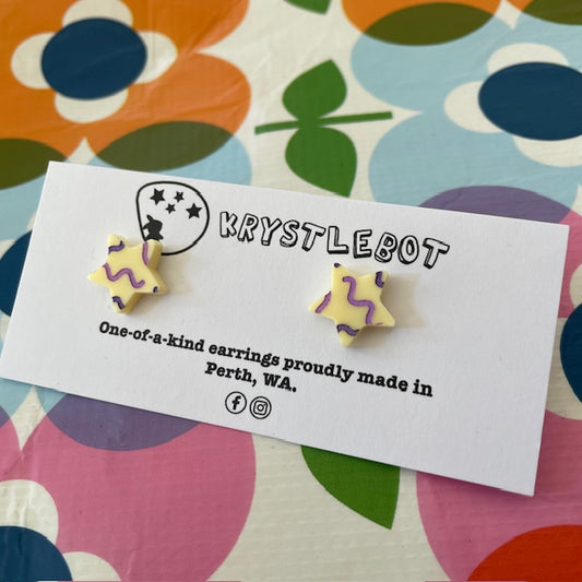 Krystlebot Squiggle Star Studs - Cream/Lilac/Purple. Star-shaped earrings with a cream coloured base and lilac and purple squiggles on a 'KRYSTLEBOT' branded card with colorful background