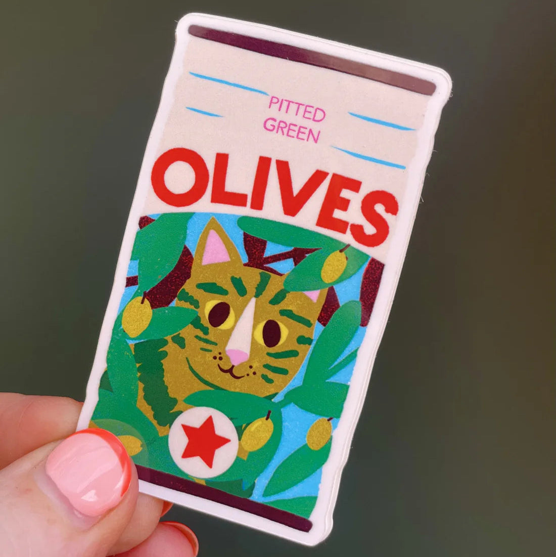 Sticker with a cat illustration labeled 'Olives' held by a hand against a dark background