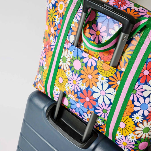 A close-up of a Project Ten Coastal Floral Medium Zip Up Tote with green and pink handles, shown resting on top of a dark grey rolling suitcase with the suitcase handle through the tote's sleeve, against a white background.