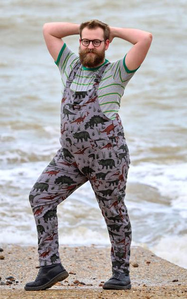 Person standing on a beach, wearing grey dungarees with a dinosaur print.