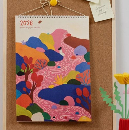 Styled image of Frankie 2026 Calendar featuring the calendar hanging on a cork notice board with a couple of notes hanging stuck next to it and a decorative flower and leaves in a vase on the bottom right of the image.