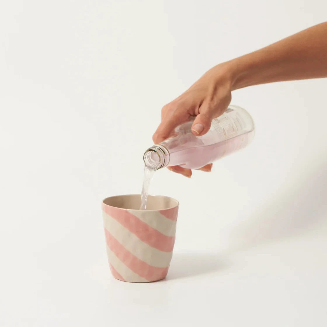 Hand pouring liquid from a bottle into a striped ceramic cup on a white background