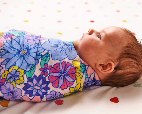 Styled image - side view of a baby swaddled in Kip & Co Bunch of Fun Bamboo Baby Swaddle on a bed with hearts on the bedsheet.