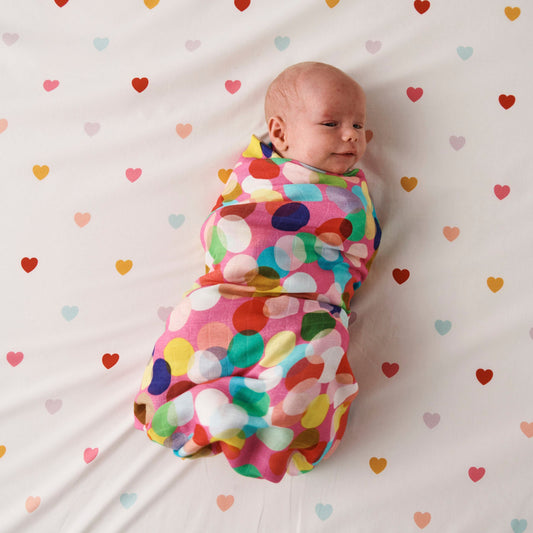 Styled image - Bird's eye view of Kip & Co Confetti Fun Bamboo Baby Swaddle swaddled around a baby on a bed with hearts on the bedsheet.