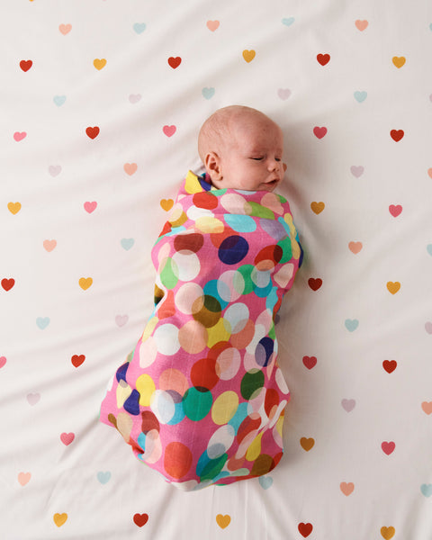 Styled image - Bird's eye view of Kip & Co Confetti Fun Bamboo Baby Swaddle swaddled around a baby on a bed with hearts on the bedsheet.