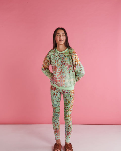 Model is standing with her hands behind her hips, looking away from the camera. She is wearing Kip&Co X Ernabella Arts Ngayaku Ngura Adult Organic Cotton Sweater. Featung artwork by Ernabella Arts artist Rupert Jack.