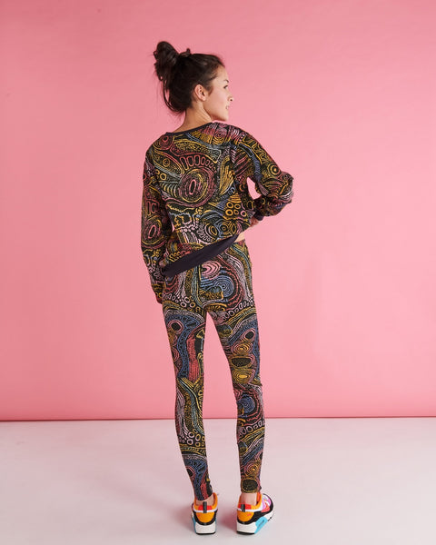 Back view of Model standing against a pastel pink backdrop with one of her hands on her hip. She is wearing a Kip&Co X Ernabella Arts Tjala Tjukurpa Adult Organic Cotton Sweater. This sweater features artwork by Ernabella Arts artist Langaliki Lewis.