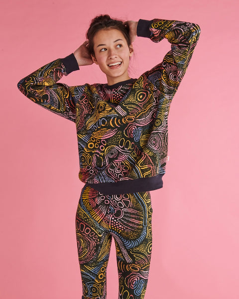 Model is standing against a pastel pink backdrop with her hands behind her head and is smiling. She is wearing a Kip&Co X Ernabella Arts Tjala Tjukurpa Adult Organic Cotton Sweater. This sweater features artwork by Ernabella Arts artist Langaliki Lewis.