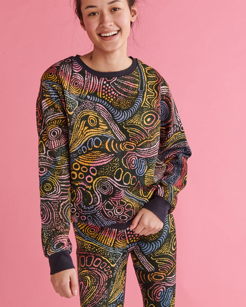 Model is standing against a pastel pink backdrop and is smiling into the camera. She is wearing a Kip&Co X Ernabella Arts Tjala Tjukurpa Adult Organic Cotton Sweater. This sweater features artwork by Ernabella Arts artist Langaliki Lewis.