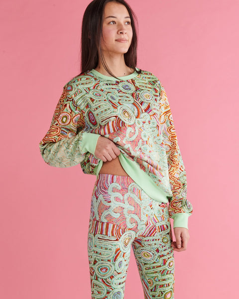 Close up of Model standing and lifting one side of the jumper slightly above the waist, and looking away from the camera. She is wearing Kip&Co X Ernabella Arts Ngayaku Ngura Adult Organic Cotton Sweater. Featung artwork by Ernabella Arts artist Rupert Jack.