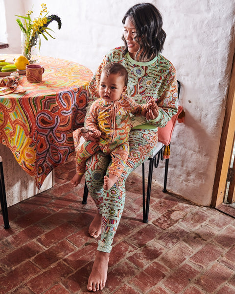 Model is sitting on a chair next to a table with an infant in her lap, looking away from the camera. She is wearing Kip&Co X Ernabella Arts Ngayaku Ngura Adult Organic Cotton Sweater. Featung artwork by Ernabella Arts artist Rupert Jack.
