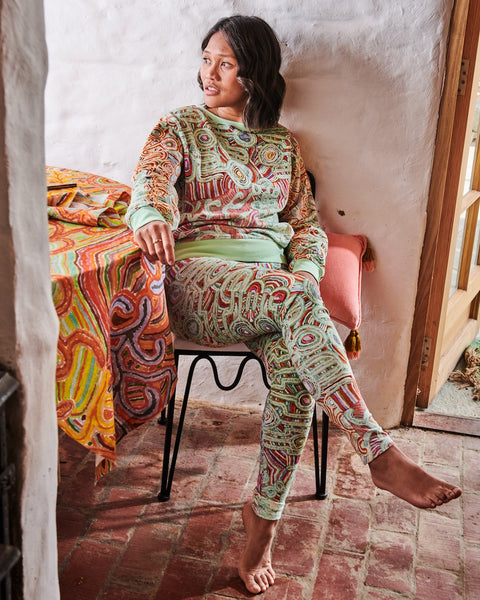 Model is sitting on a chair next to a table with a patterned tablecloth. She is wearing Kip&Co X Ernabella Arts Ngayaku Ngura Adult Organic Cotton Sweater. Featung artwork by Ernabella Arts artist Rupert Jack.