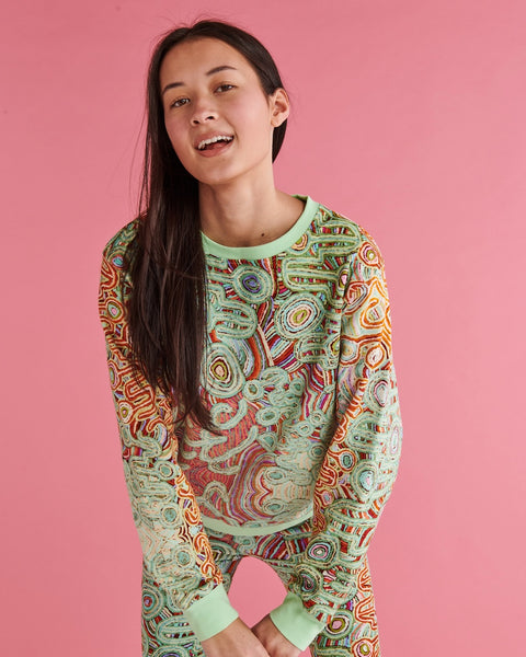 Model is standing slouched forward with her hands in front of her, looking at the camera. She is wearing Kip&Co X Ernabella Arts Ngayaku Ngura Adult Organic Cotton Sweater. Featung artwork by Ernabella Arts artist Rupert Jack.
