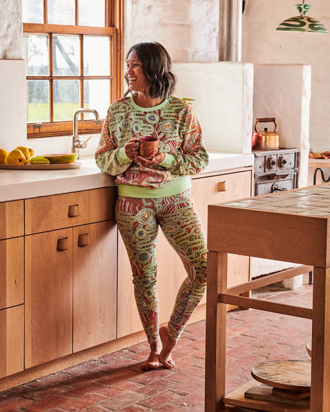 Model is in a kitchen casually leaning against the counter next to the sink, and holding a mug. She is wearing Kip&Co X Ernabella Arts Ngayaku Ngura Adult Organic Cotton Sweater. Featung artwork by Ernabella Arts artist Rupert Jack.