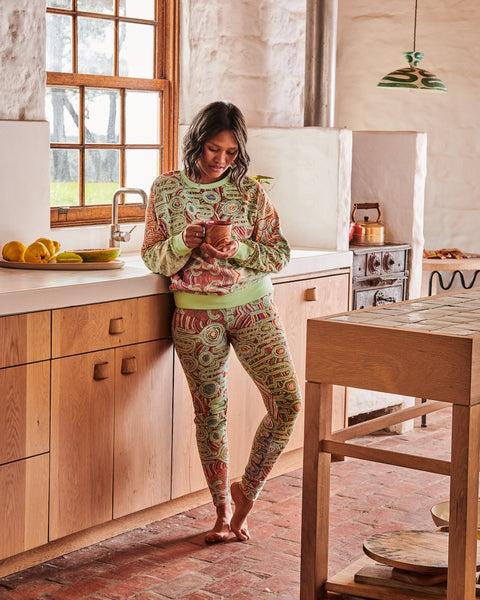 Model is standing against a kitchen counter holding a mug with both hands and looking down at the mug. She is wearing Kip&Co X Ernabella Arts Ngayaku Ngura Adult Organic Cotton Sweater. Featung artwork by Ernabella Arts artist Rupert Jack. 