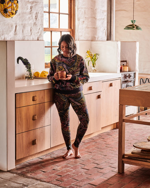 Model is leaning against the kitchen counter holding a mug with both hands, looking down at the mug. She is wearing a Kip&Co X Ernabella Arts Tjala Tjukurpa Adult Organic Cotton Sweater. This sweater features artwork by Ernabella Arts artist Langaliki Lewis.