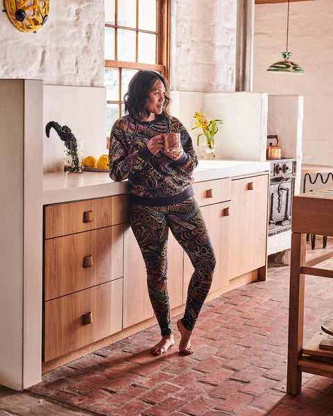 Model is leaning against the kitchen counter holding a mug with both hands, looking away from the camera. She is wearing a Kip&Co X Ernabella Arts Tjala Tjukurpa Adult Organic Cotton Sweater. This sweater features artwork by Ernabella Arts artist Langaliki Lewis.