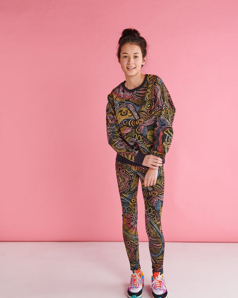 Model is standing against a pastel pink backdrop with her hands clasped in front of her, and is smiling at the camera. She is wearing a Kip&Co X Ernabella Arts Tjala Tjukurpa Adult Organic Cotton Sweater. This sweater features artwork by Ernabella Arts artist Langaliki Lewis.