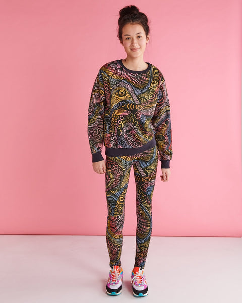 Model is standing against a pastel pink backdrop and is smiling. She is wearing a Kip&Co X Ernabella Arts Tjala Tjukurpa Adult Organic Cotton Sweater. This sweater features artwork by Ernabella Arts artist Langaliki Lewis.