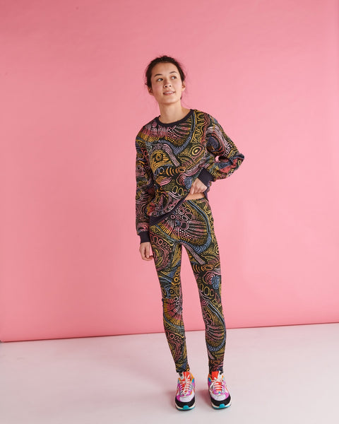 Model is standing against a pastel pink backdrop with one of her hands lifting a side of the jumper above her waist, and is smiling away from the camera. She is wearing a Kip&Co X Ernabella Arts Tjala Tjukurpa Adult Organic Cotton Sweater. This sweater features artwork by Ernabella Arts artist Langaliki Lewis.