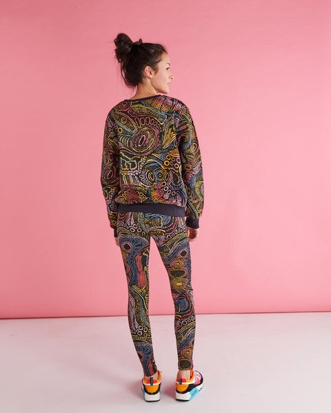 Back view of Model standing against a pastel pink backdrop. She is wearing a Kip&Co X Ernabella Arts Tjala Tjukurpa Adult Organic Cotton Sweater. This sweater features artwork by Ernabella Arts artist Langaliki Lewis.