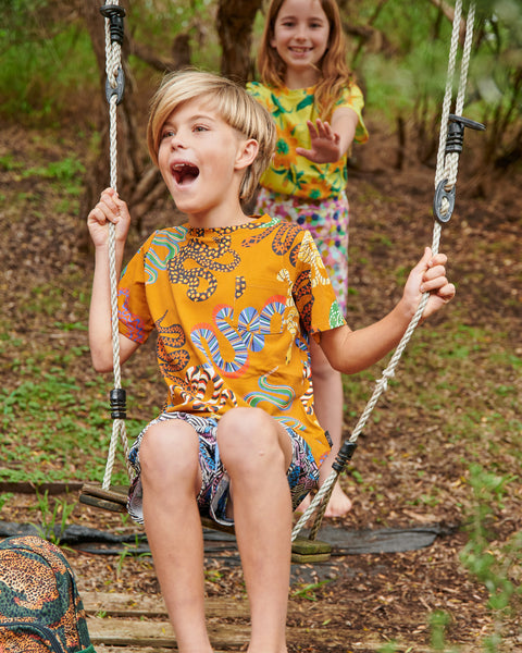 Kip & Co Snake Pit Kid's Tee worn by a young model sitting on a swing being pushed by another young model in the background