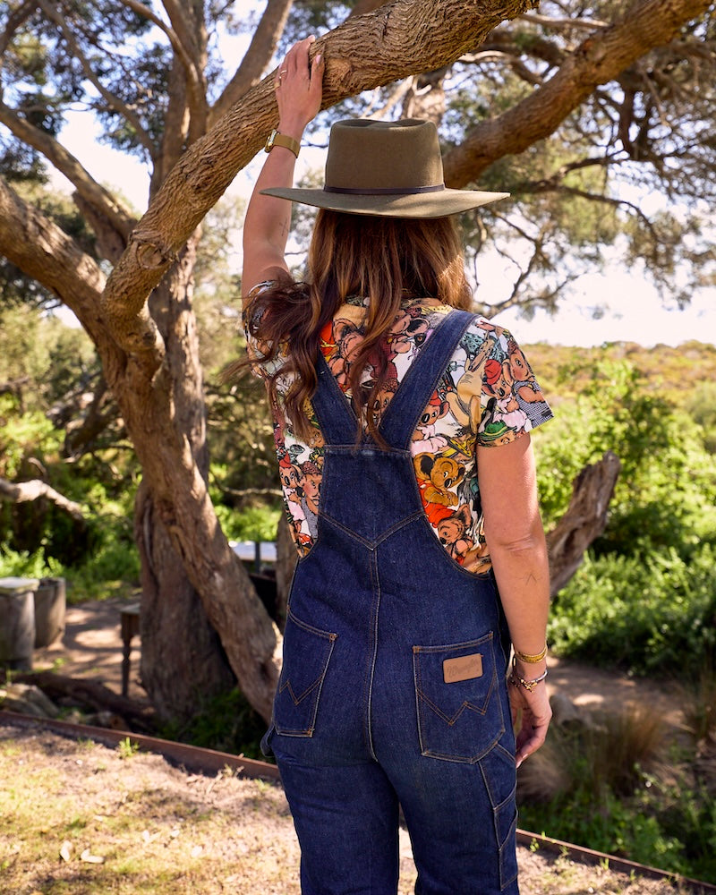 A woman viewed from behind wearing blue denim overalls over a colourful Blinky Bill short-sleeve shirt, a wide-brimmed brown hat, and bracelets, standing outdoors under a tree.