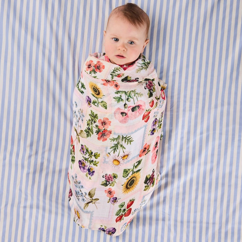 A baby swaddled in a light pink muslin swaddle blanket with a colorful floral print, lying on a blue and white striped sheet.
