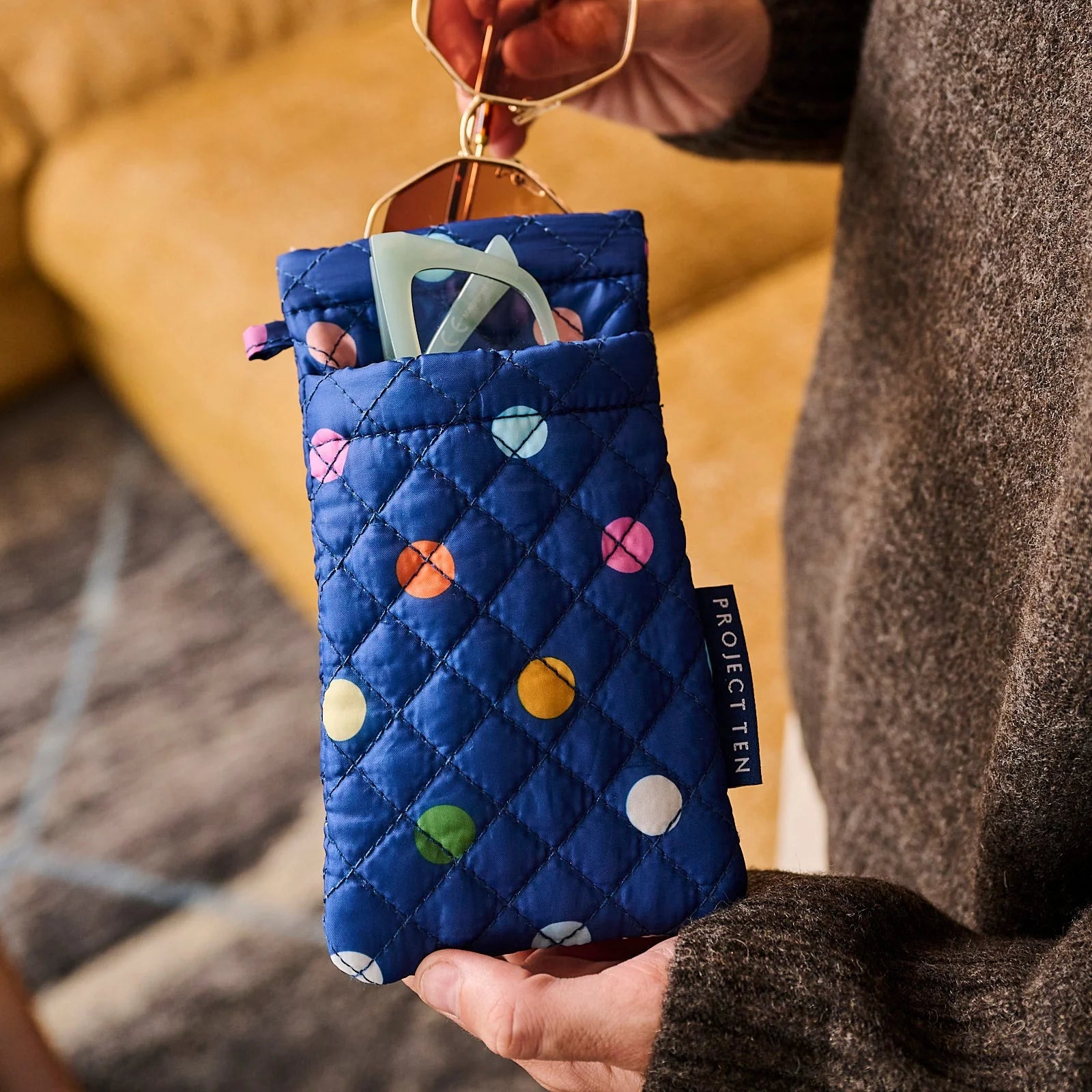 Styled image of Blue quilted pouch with colorful polka dots holding sunglasses, held by a person.