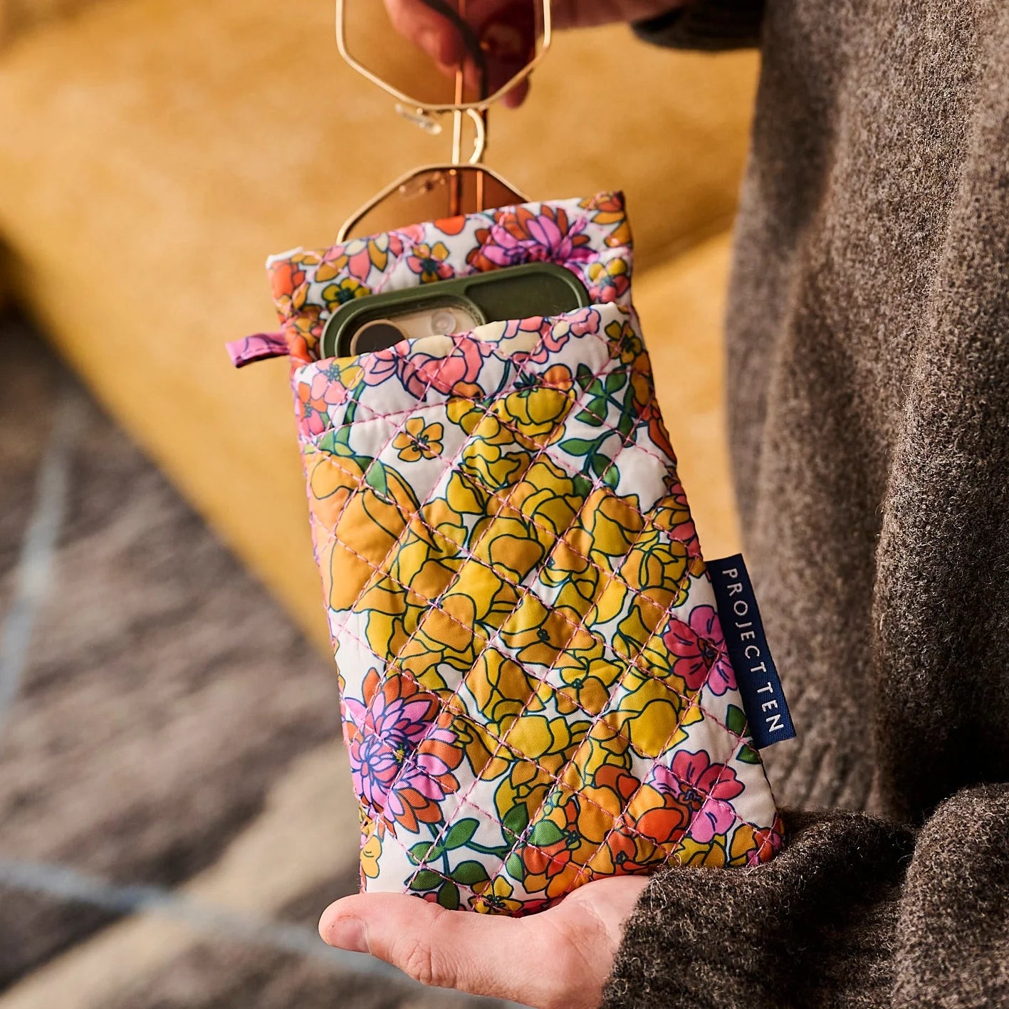 Floral patterned glasses case with sunglasses peeking from top of case and a mobile phone in the front pocket held by a person wearing a brown jumper.  