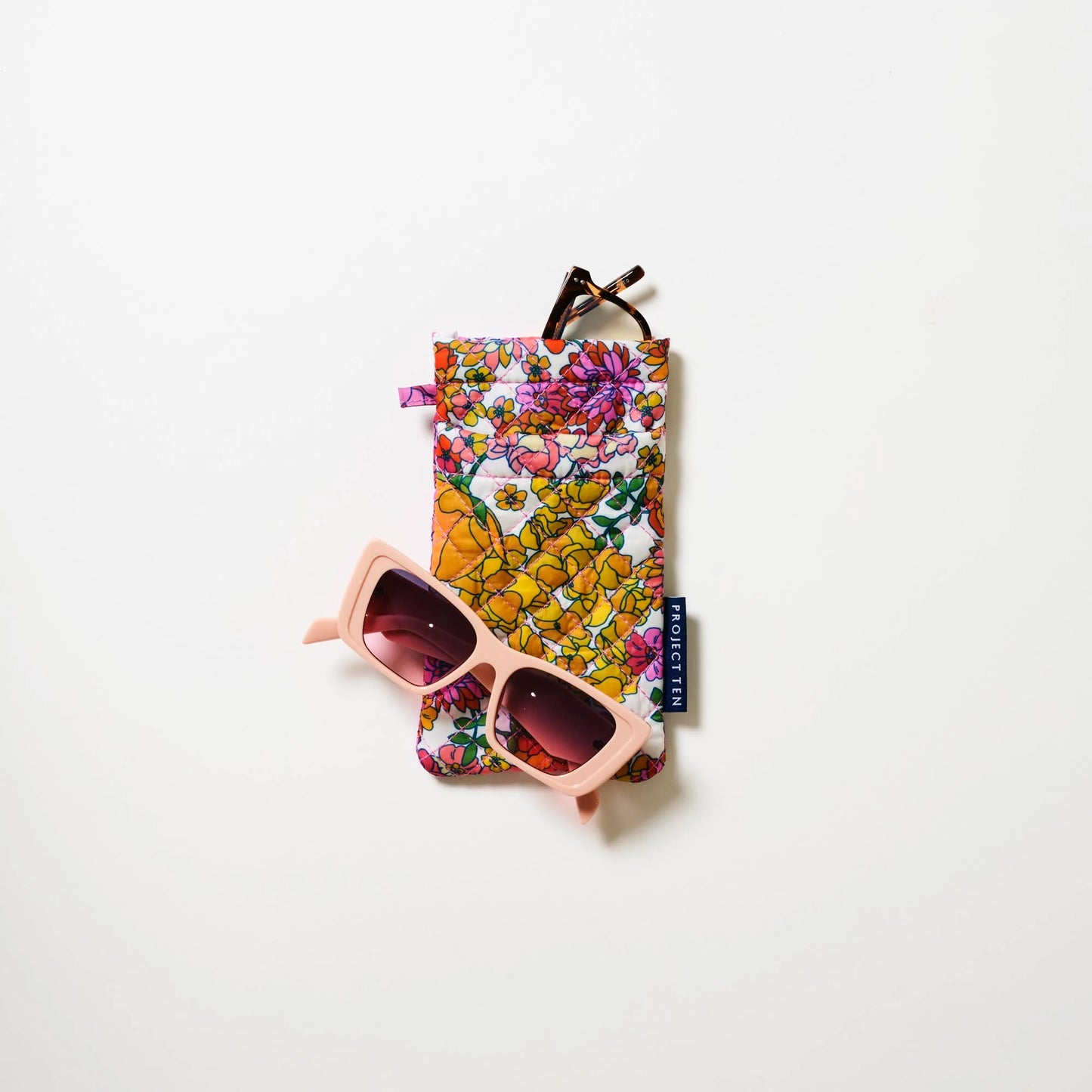 Styled image of Floral sunglasses case with pink sunglasses resting on the front of case and a pair of reading glasses peeking from the top of case on a white background