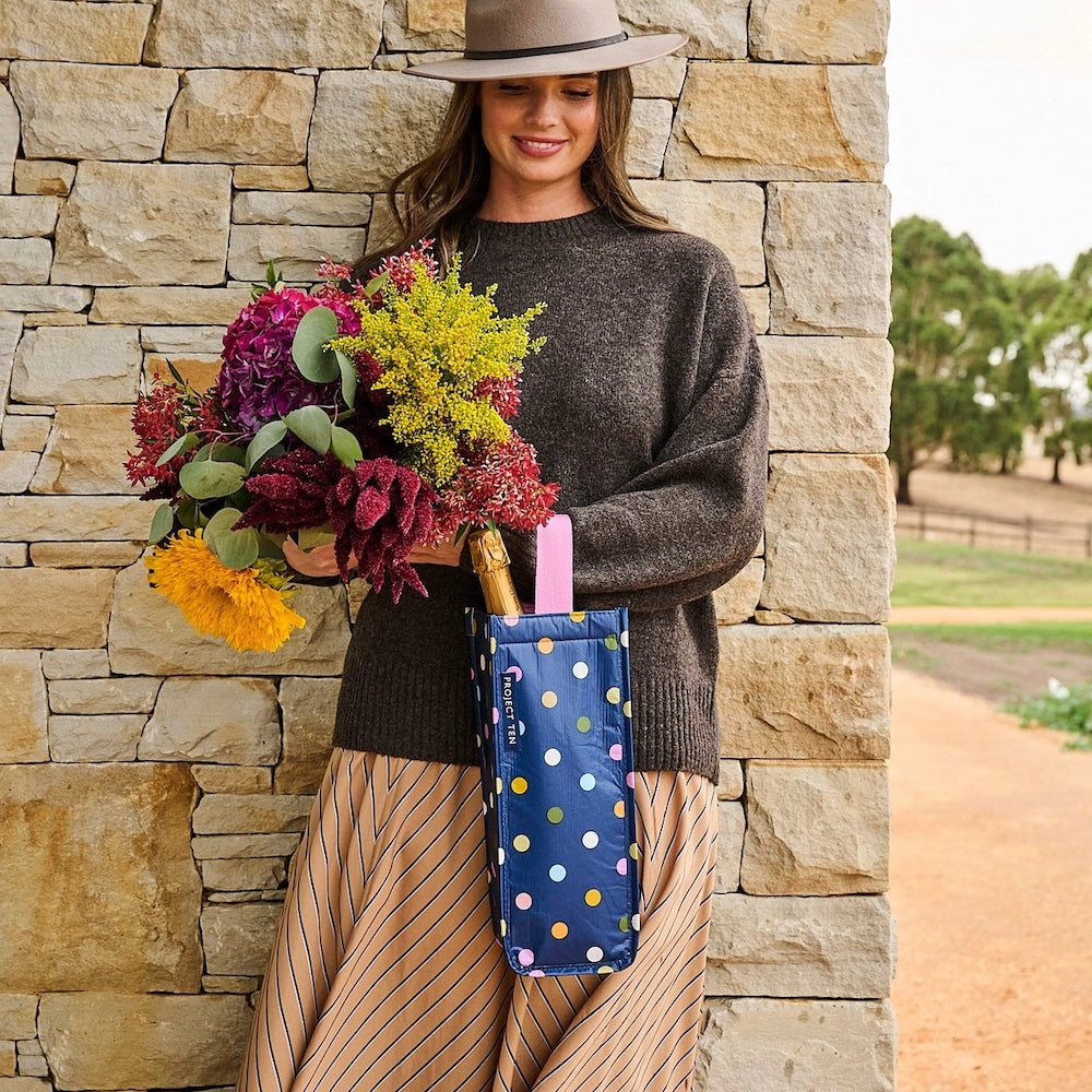 Woman holding a bouquet of flowers and a polka dot bag against a stone wall.