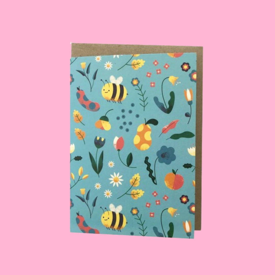 A greeting card with a colorful nature-themed pattern of bees, flowers, leaves, and acorns on a light blue background, shown with a brown kraft envelope against a solid pink backdrop.