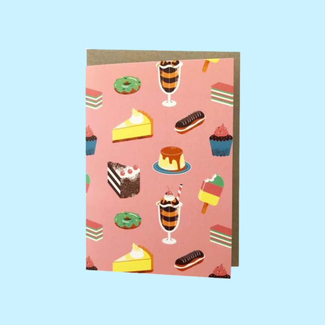 A pink greeting card featuring a repeating pattern of illustrated desserts, including cakes, cupcakes, ice cream, and donuts, shown with a brown envelope against a light blue background.