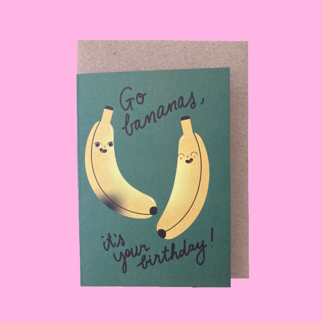 A birthday greeting card featuring two smiling illustrated bananas on a dark green background with the text 'Go bananas, it's your birthday!', shown with a kraft envelope against a solid pink background.