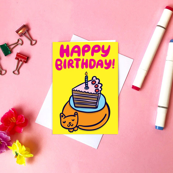 Styled image of Able & Game Cake on Cat Happy Birthday Card surrounded by a couple of pens, bull dog clips and a few plastic flowers