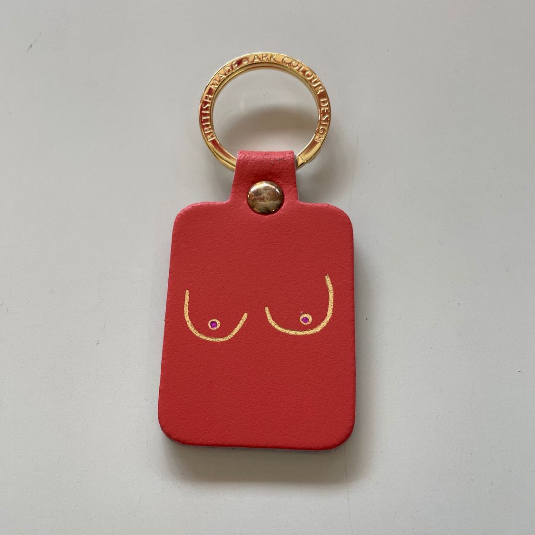 A coral-red leather keychain tag featuring a minimalist gold line-art design of two breasts with small purple dots for nipples, attached to a gold metal keyring.