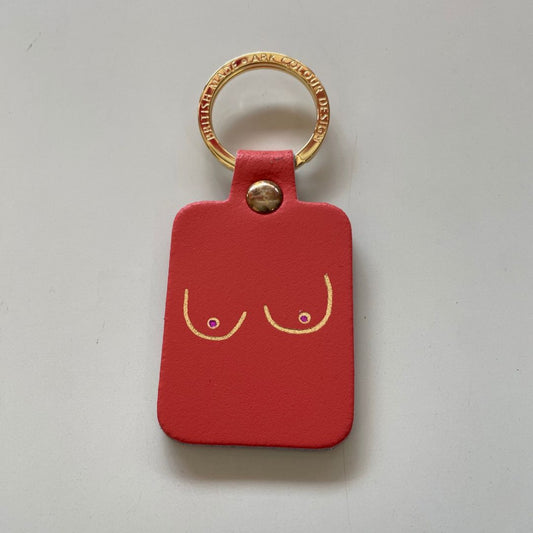 A coral-red leather keychain tag featuring a minimalist gold line-art design of two breasts with small purple dots for nipples, attached to a gold metal keyring.
