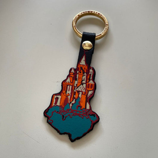 A leather keychain shaped like a castle on clouds, featuring a metallic orange foil and white castle, a teal foreground looking like clouds, and a gold-tone metal key ring and rivet.