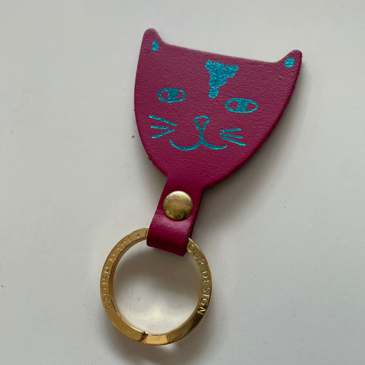 A magenta leather keychain shaped like a cat head with turquoise foil facial features and a gold-tone split ring, shown on a plain light background.